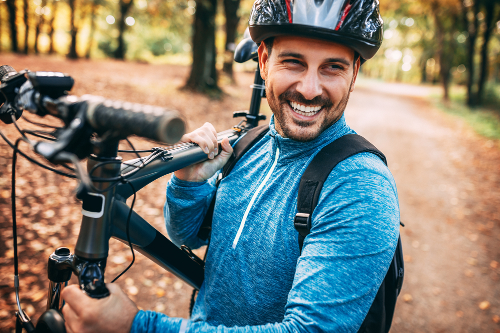 YA man holds his mountain bike with his helmet on and smiles on a trail